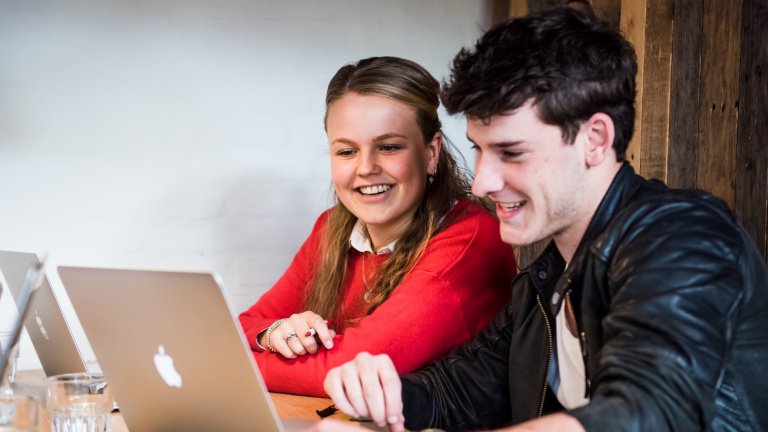Twee hotelschool studenten zitten lachend achter hun Apple laptop te studeren in een van de rode cabines op de 1e verdieping van het studentenrestaurant Refresh