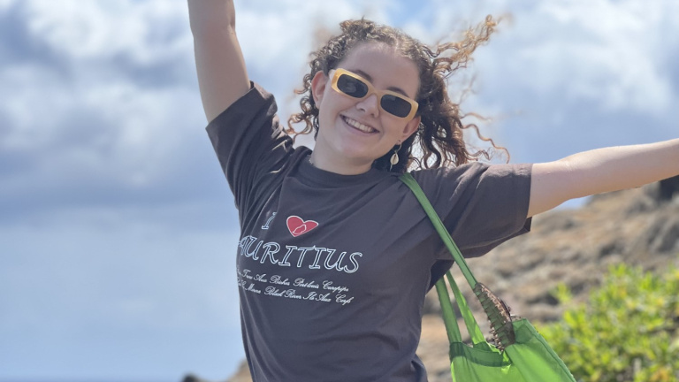girl with curls and yellow sunglasses, wearing a brown shirt 'i love mauritius', sitting on a rock near the sea