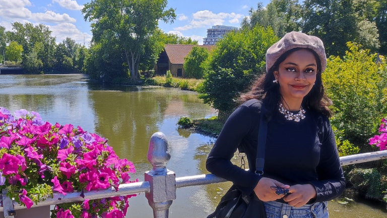 Girl with dark hair, black top and jeans, standing on a bridge in front of water. Pink Flowers and Green Trees in Background