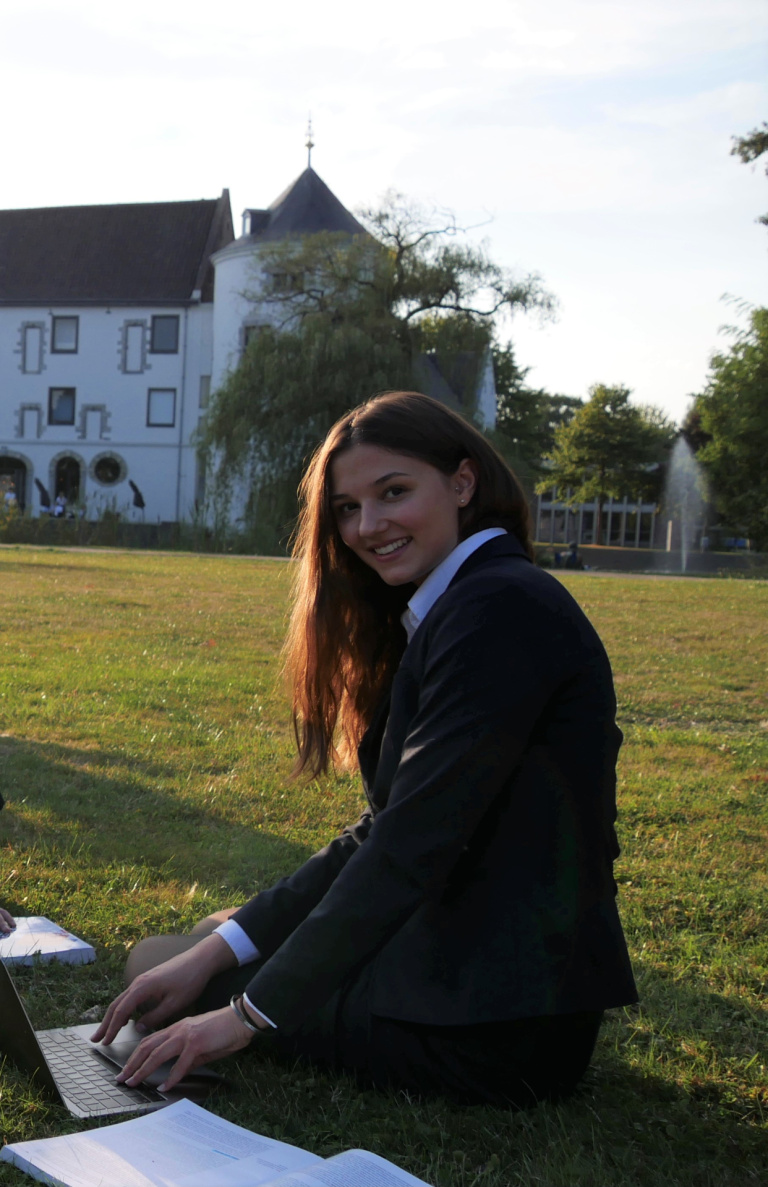 A student sitting in the grass in front of the Teaching Hotel. She is working on her laptop.