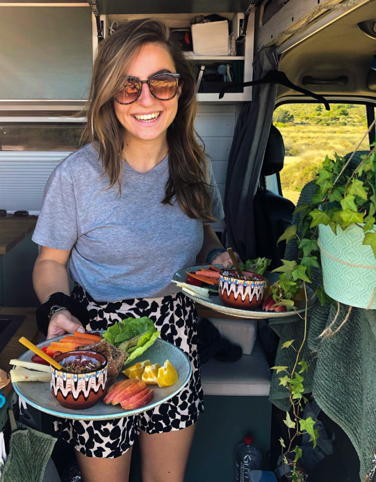Meisje met donkerbruin haar en een zonnebril op, grijs shirt, rokje, met fruit in haar handen in een busje